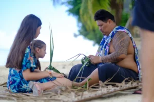 Un homme Polynésien montre comment réaliser un tressage de palme de cocotier à deux enfants assis sur la plage du Tiki Village en face de lui.