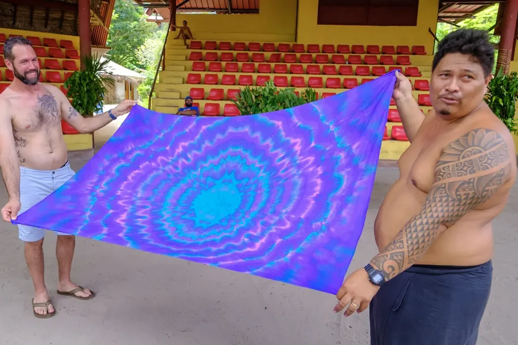 Deux homme tiennent un paréo coloré tendu entre eux après un atelier de confection de Paréo. Ils sont au milieu de la scène du Tiki Village ° Moorea