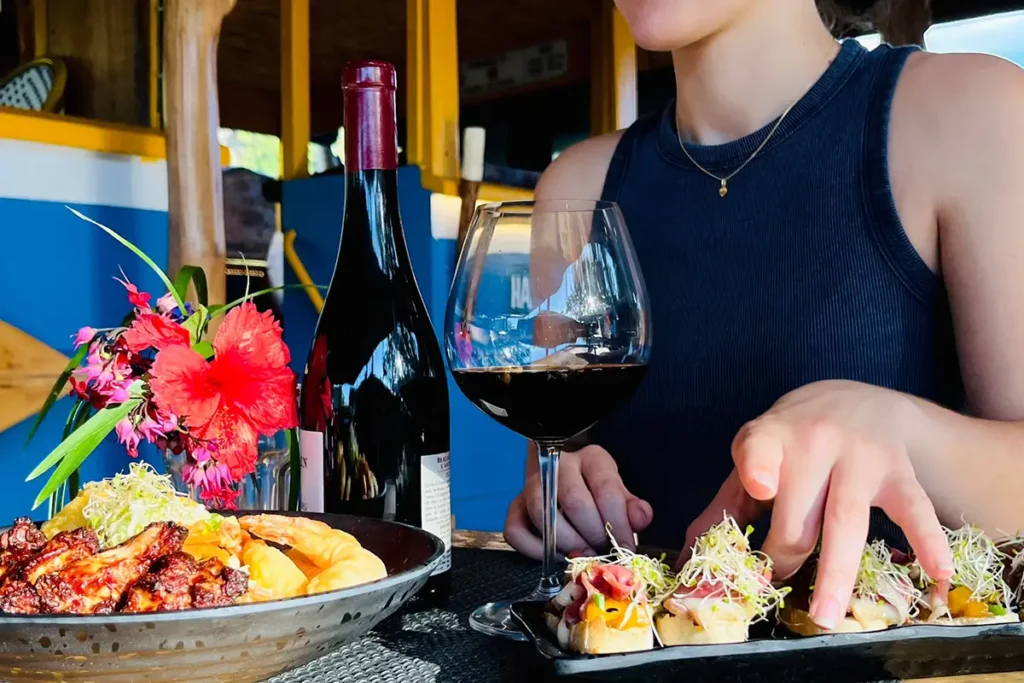 Gros plan sur le buste d'une femme qui se tient derrière une table de restaurant avec devant elle des assiettes de plats polynésiens et une bouteille de vin rouge ainsi qu'un verre à pied. Scene du restaurant Truck Uaina à Moorea
