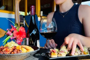 Gros plan sur le buste d'une femme qui se tient derrière une table de restaurant avec devant elle des assiettes de plats polynésiens et une bouteille de vin rouge ainsi qu'un verre à pied. Scene du restaurant Truck Uaina à Moorea