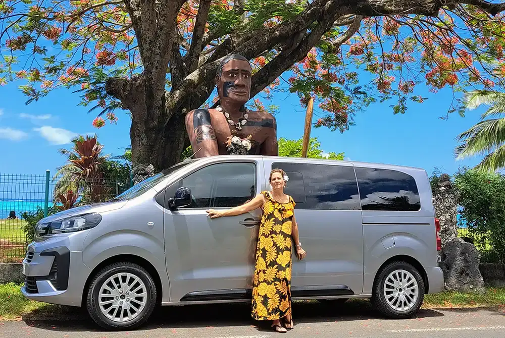 Une femme habillée en longue robe tahitienne se tient devant une voiture van de transport privé à Moorea. En arrière plan, un arbre flamboyant et une grande statue polynésienne agenouillée sous l'arbre
