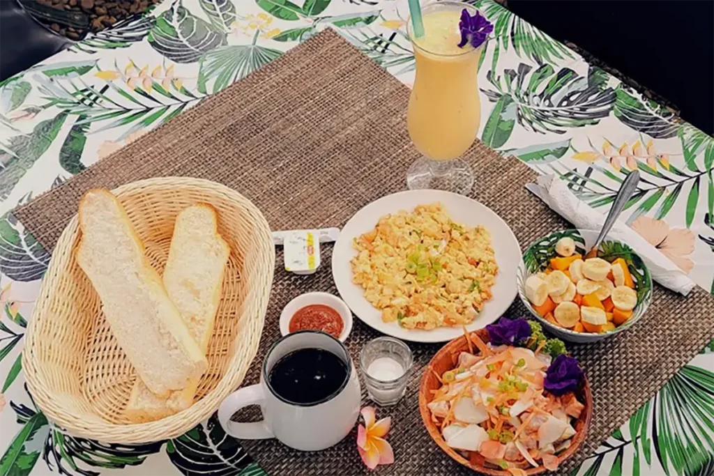 Une table de petit déjeuner traditionnel Tahitien vue de haut, avec une tasse de café, du pain, un smoothie, des oeufs brouillés, une salade de fruit et du poisson cru à la tahitienne