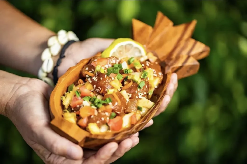Les mains d'un homme tiennent un petit plat creux en bois en forme d'ananas dans lequel est servi un poke de poisson frais à la tahitienne