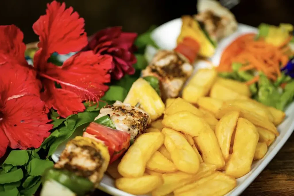 Une assiette blanche rectangulaire dans laquelle est posé une brochette de poisson grillé servi avec des frites et des légumes. Des fleurs d'hibiscus rouges sont posées sur le coté gauche de l'assiette