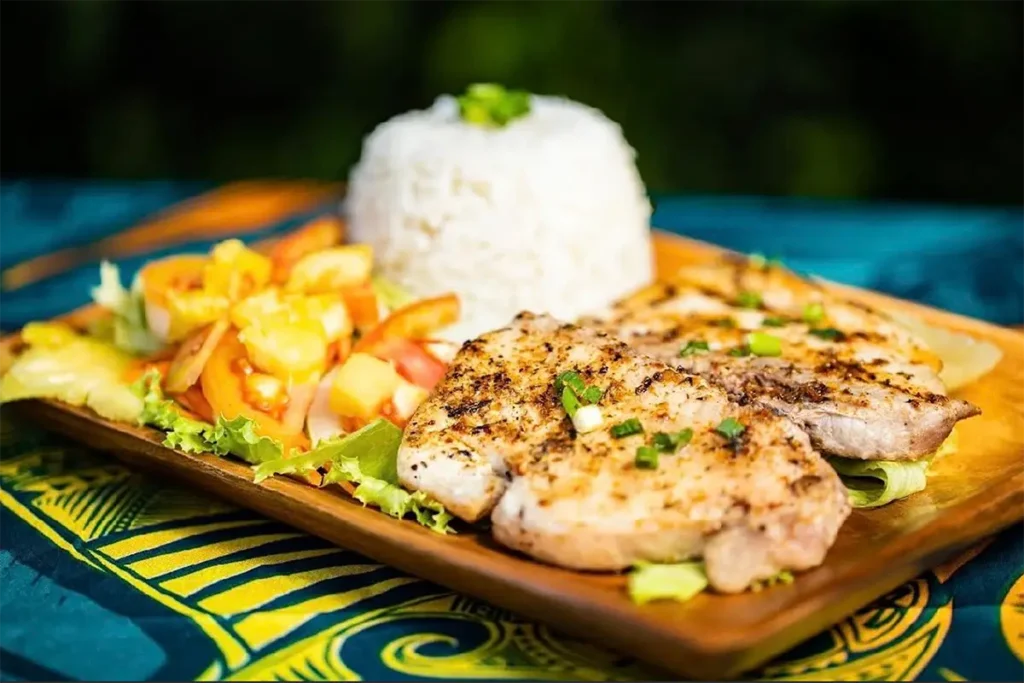 Une assiette rectangulaire en bois avec du poisson grillé de Polynésie, du riz et des légumes. L'assiette est posée sur un pareo Tahitien bleu et jaune