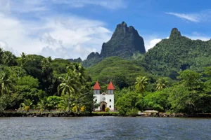 Sainte-Famille Church in Haapiti seen from the lagoon with palm trees and a shark-tooth-shaped volcanic peak in Moorea