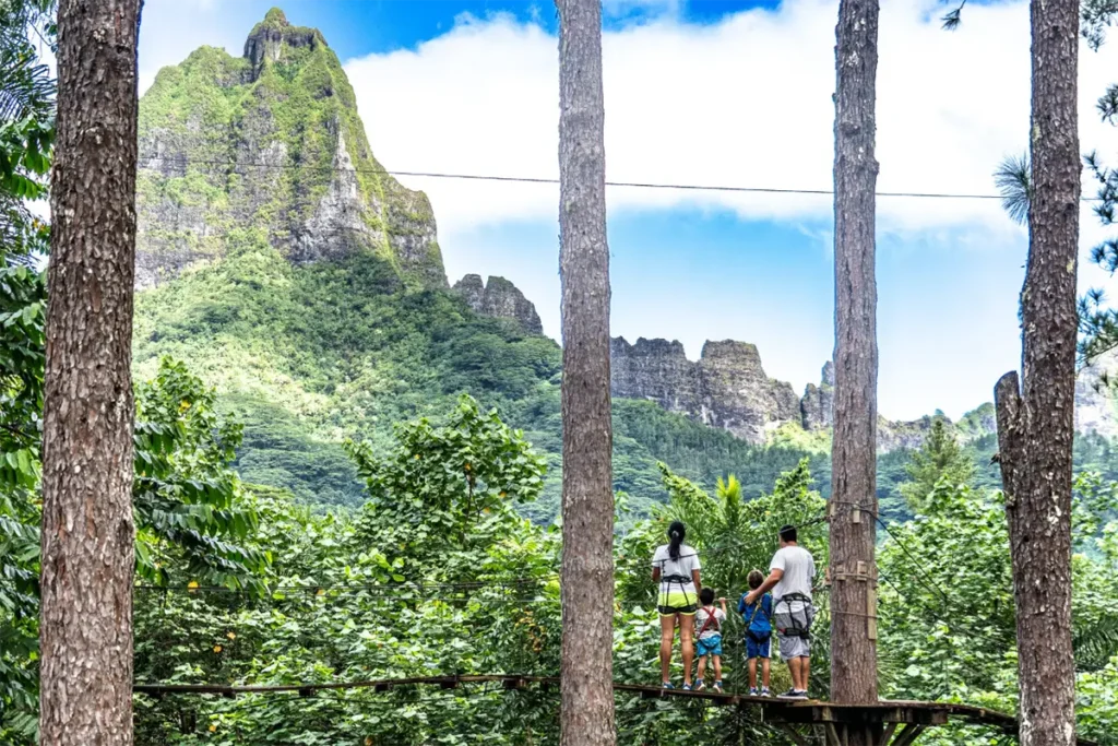 Une famille se tient de dos sur une plateforme en bois sur une étape du parcours aventure du tiki parc de Moorea. Ils regarde la paysage de montagne en arrière plan