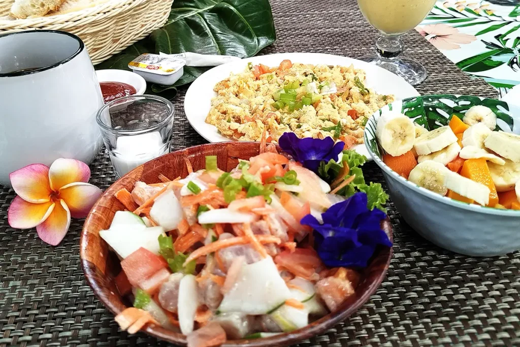 Vue d'ensemble d'une table de petit déjeuner dans un restaurant à Moorea. Des plats y sont présentés: poisson cru, salade de fruit, œufs brouillés et tasse de café.