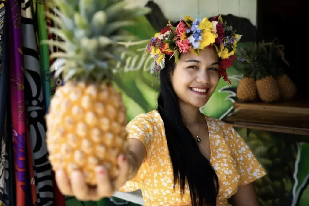 Une jeune femme tahitienne avec une couronne de fleur sur la tête tient devant elle bras tendu, un ananas