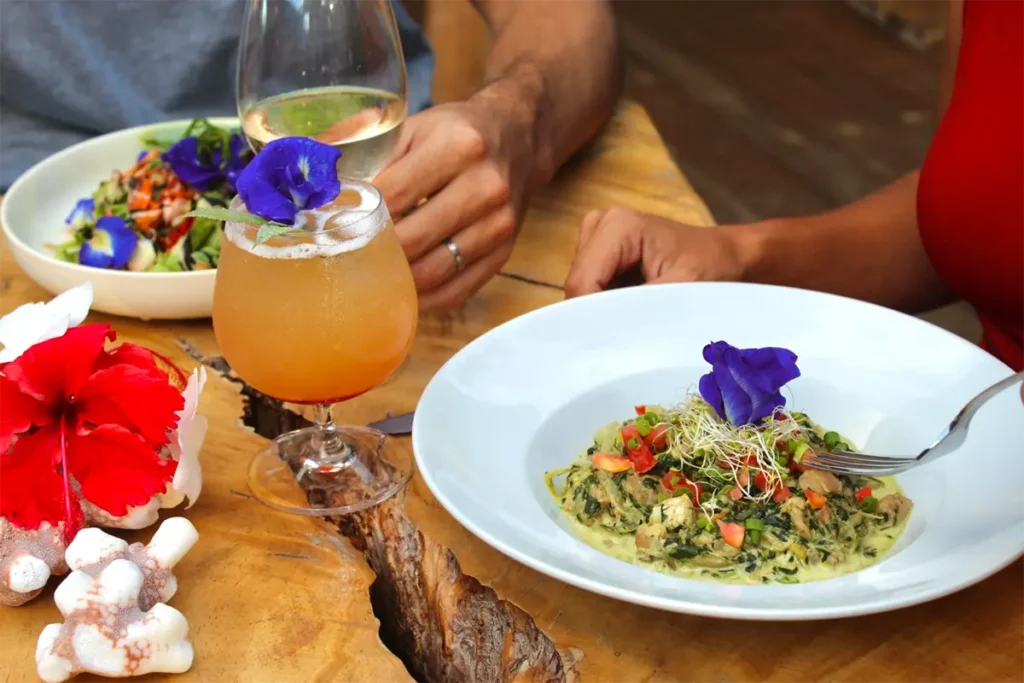 Table d'un restaurant Tahitien à Moorea. Un verre de cocktail est posé au centre et gros plan sur deux assiettes servies avec un repas local de légumes. Une fleur d'hibiscus rouge sur la gauche de la table