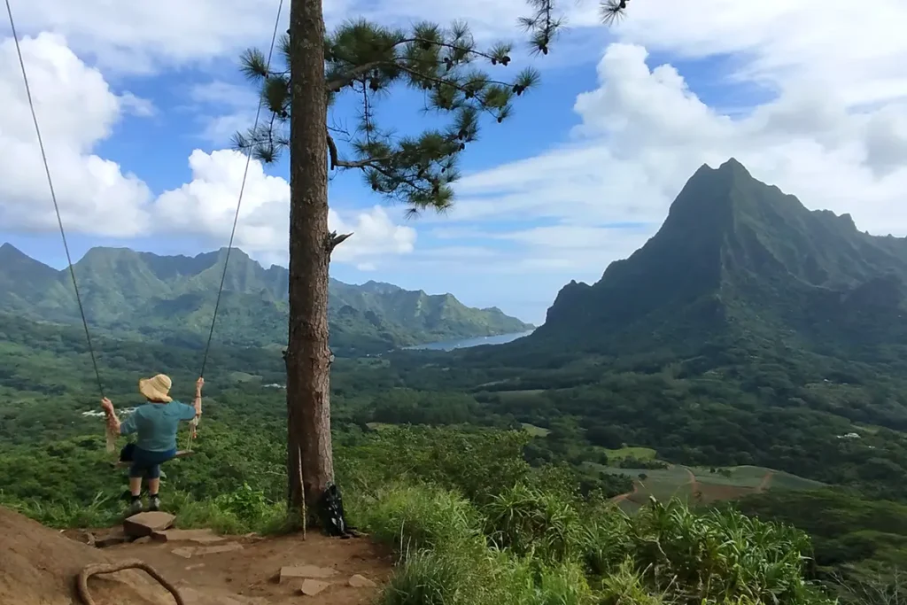 Une vielle femme avec un chapeau tressé sur la tête, est assise sur un balançoire devant un panorama de montagnes de Moorea.