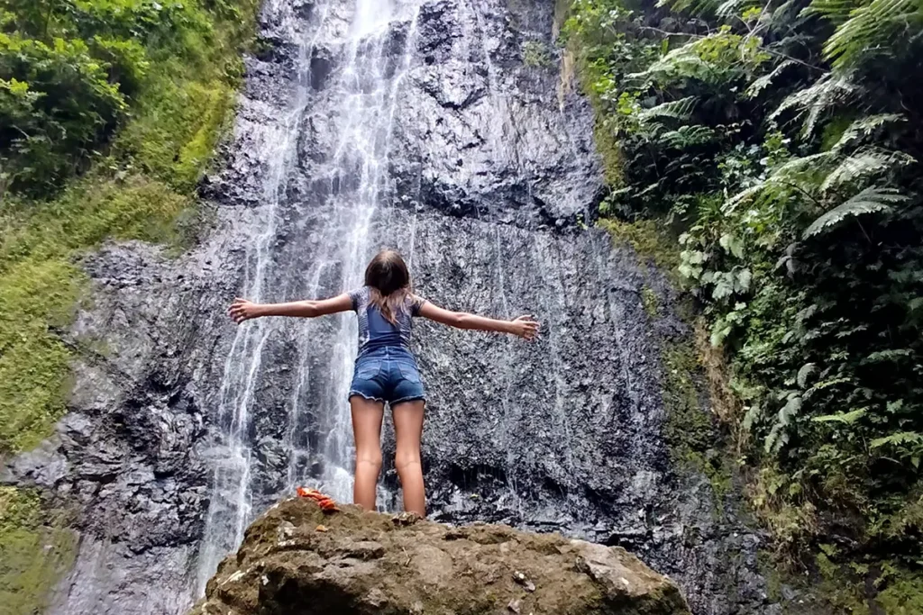 Une jeune fille se tient les bras en croix devant la cascade de Afareaitu à Moorea. Elle regarde en l'air.